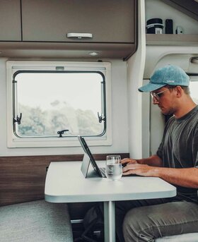 Un uomo lavora in un camper Carado nella dinette con tavolo, tablet e finestra all'interno.