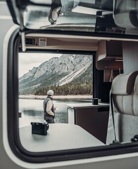 Vista dal camper Carado di un uomo in riva al lago con paesaggio montano sullo sfondo; interno con tavolo e cucina