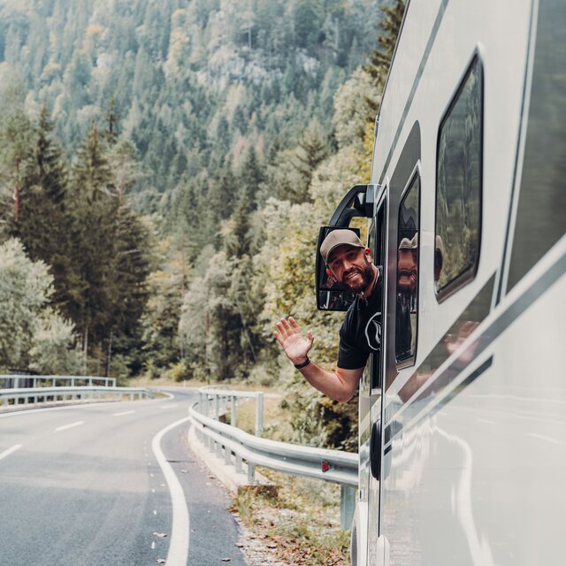 A man looks out from a Carado motorhome and waves on a winding mountain road at the edge of the forest.