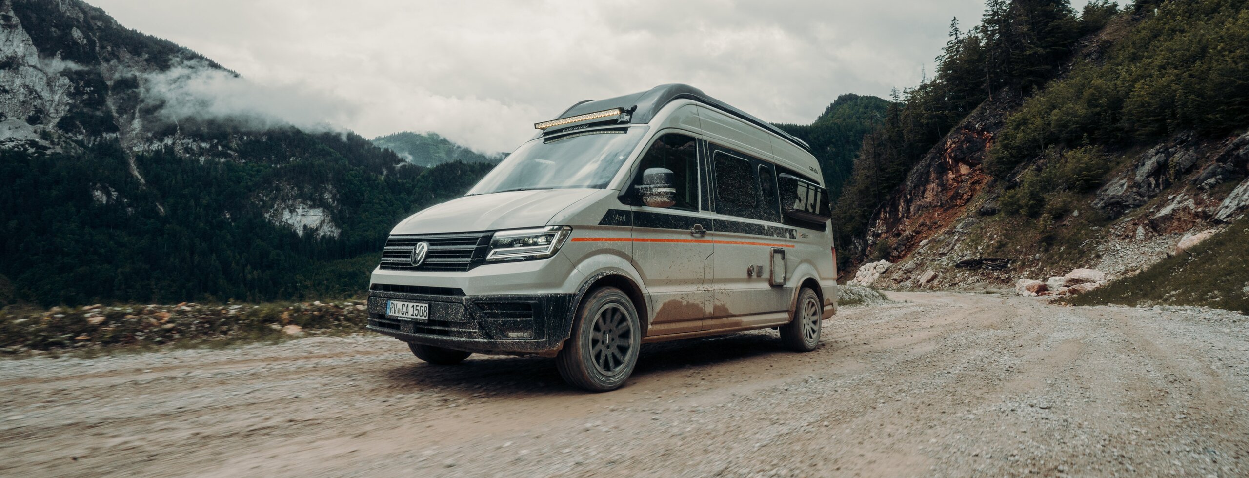 Carado CV595 4x4 X-EDITION fährt über eine Schotterpiste vor einem Bergmassiv