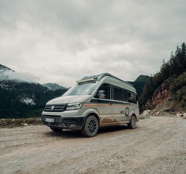 Carado CV595 4x4 X-EDITION fährt über eine Schotterpiste vor einem Bergmassiv
