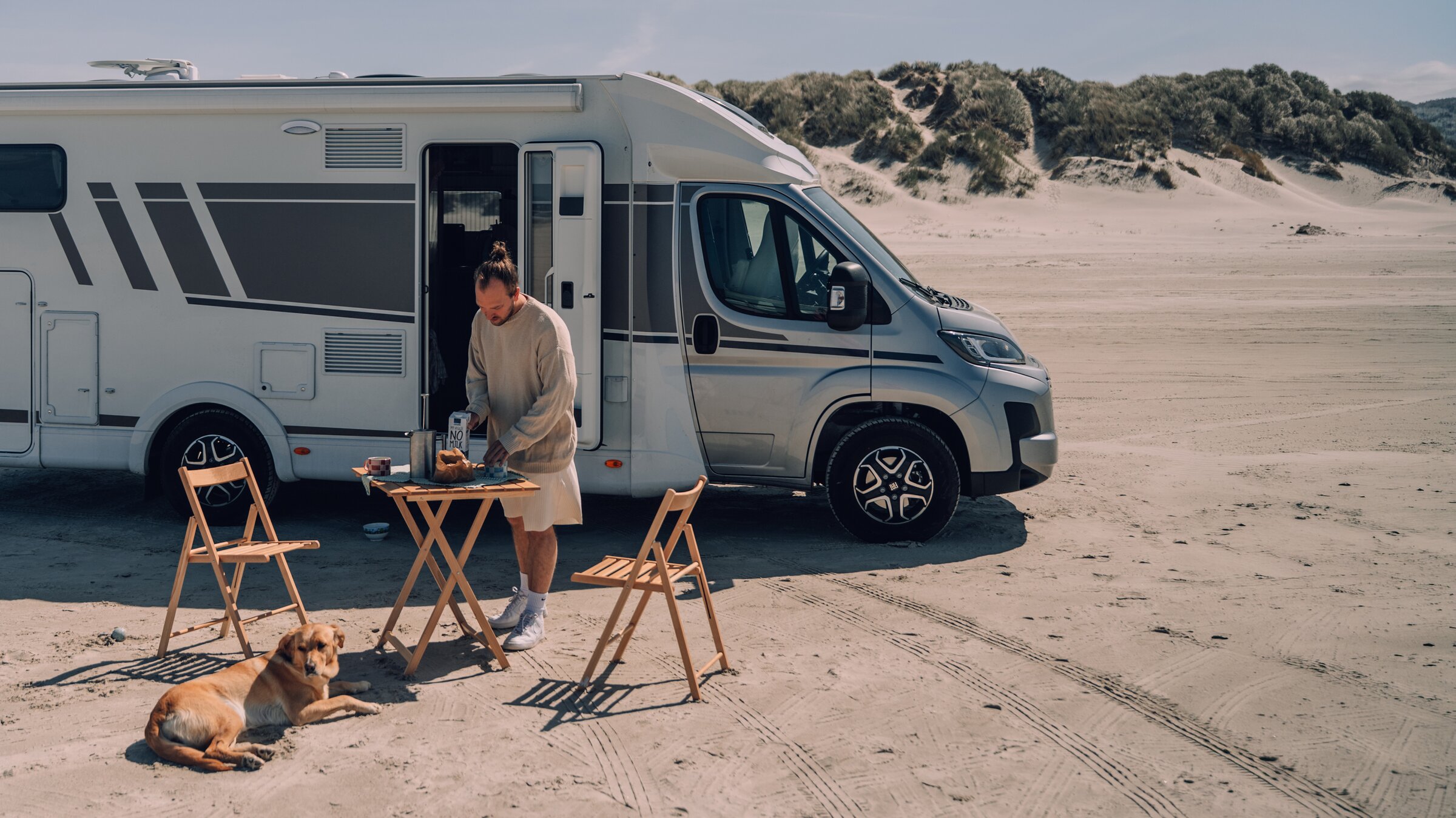 Mann bereitet das Frühstück an einem kleinen Holztisch vor einem Wohnmobil direkt am Sandstrand zu, daneben liegt ein Hund im Sand.