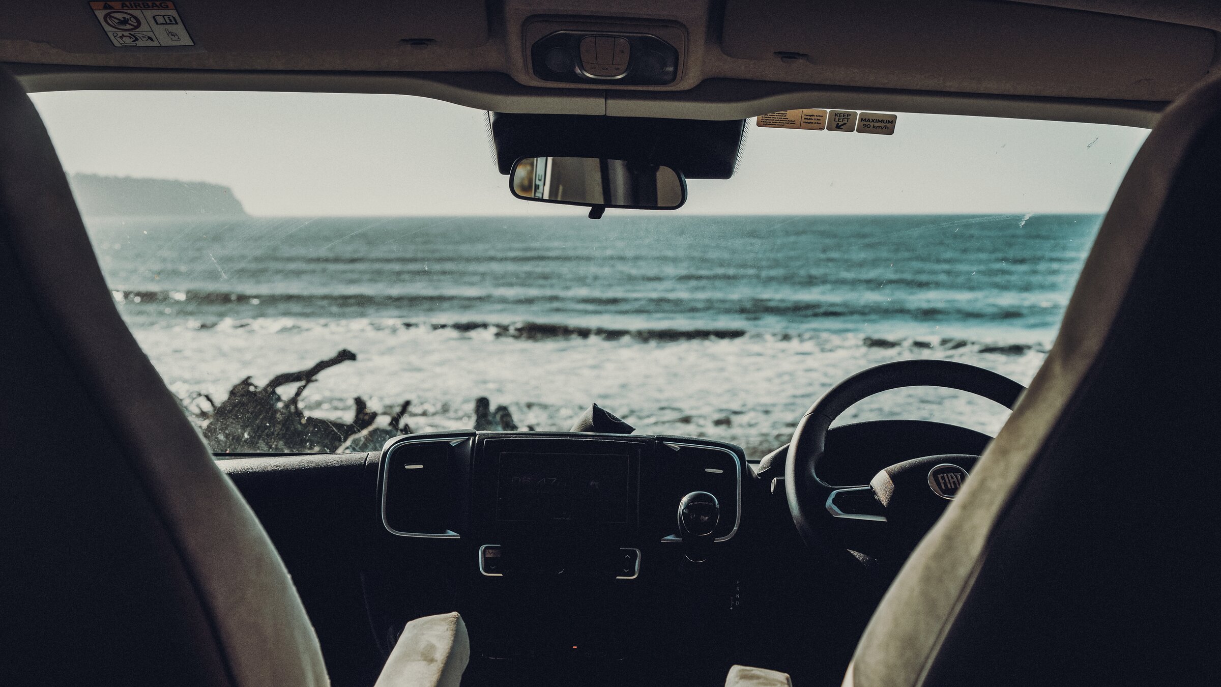 Blick aus dem Cockpit eines geparkten Campervans durch die Windschutzscheibe auf den Strand und das Meer.