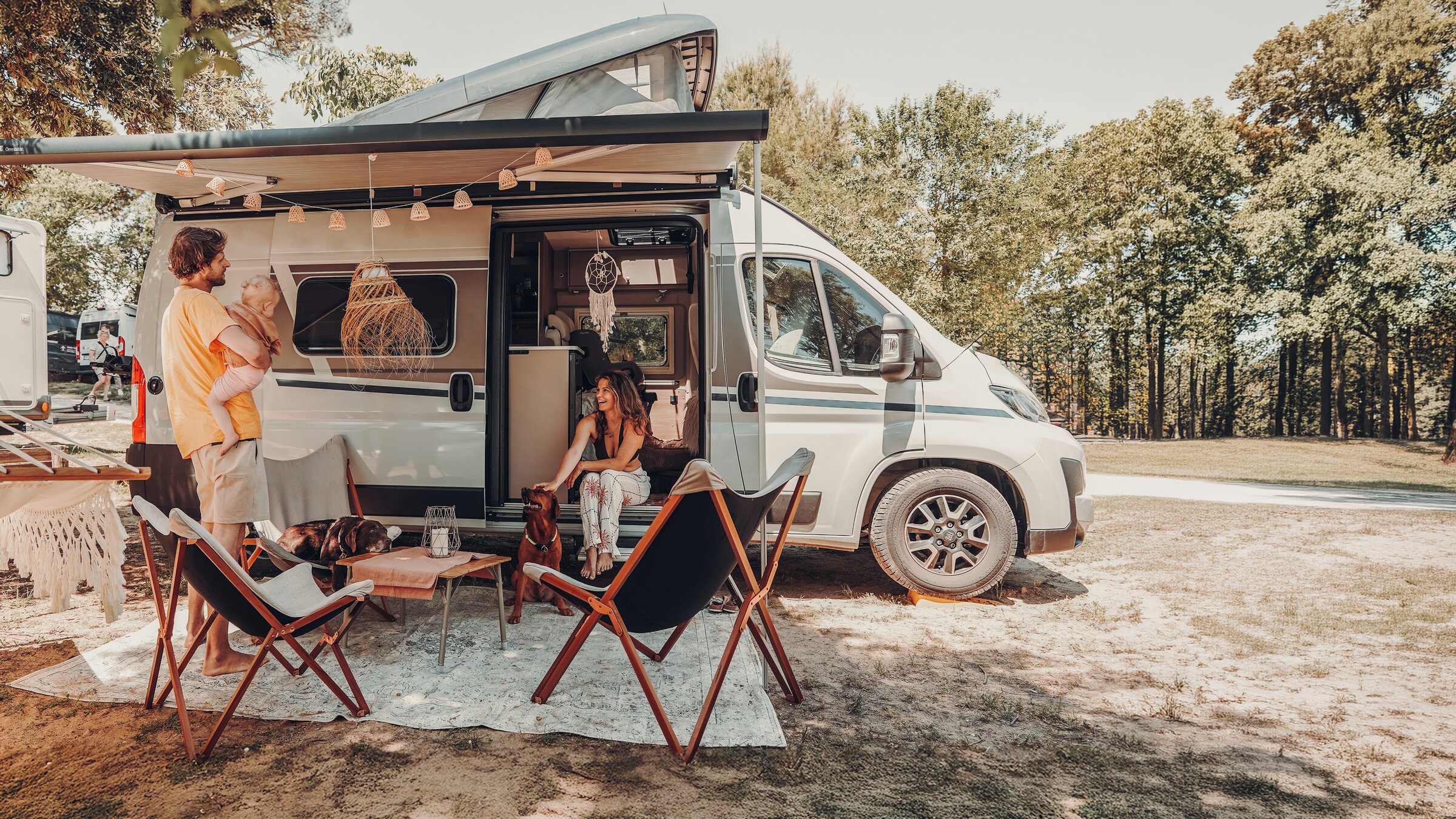Junge Familie mit Baby und Hund entspannt unter der Markise eines Campervans auf einem sonnigen Stellplatz im Grünen.