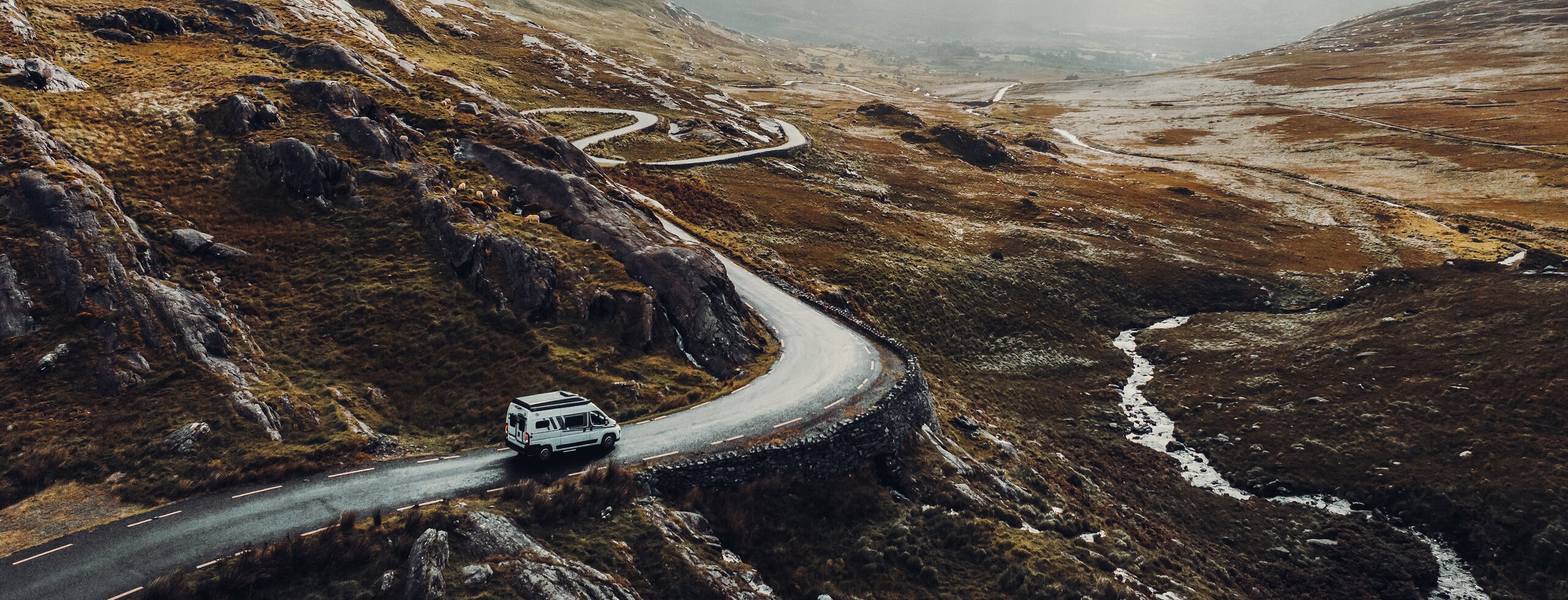 Ein Campervan fährt auf einer einsamen Bergstraße durch eine raue, steinige Gebirgslandschaft bei bewölktem Himmel.