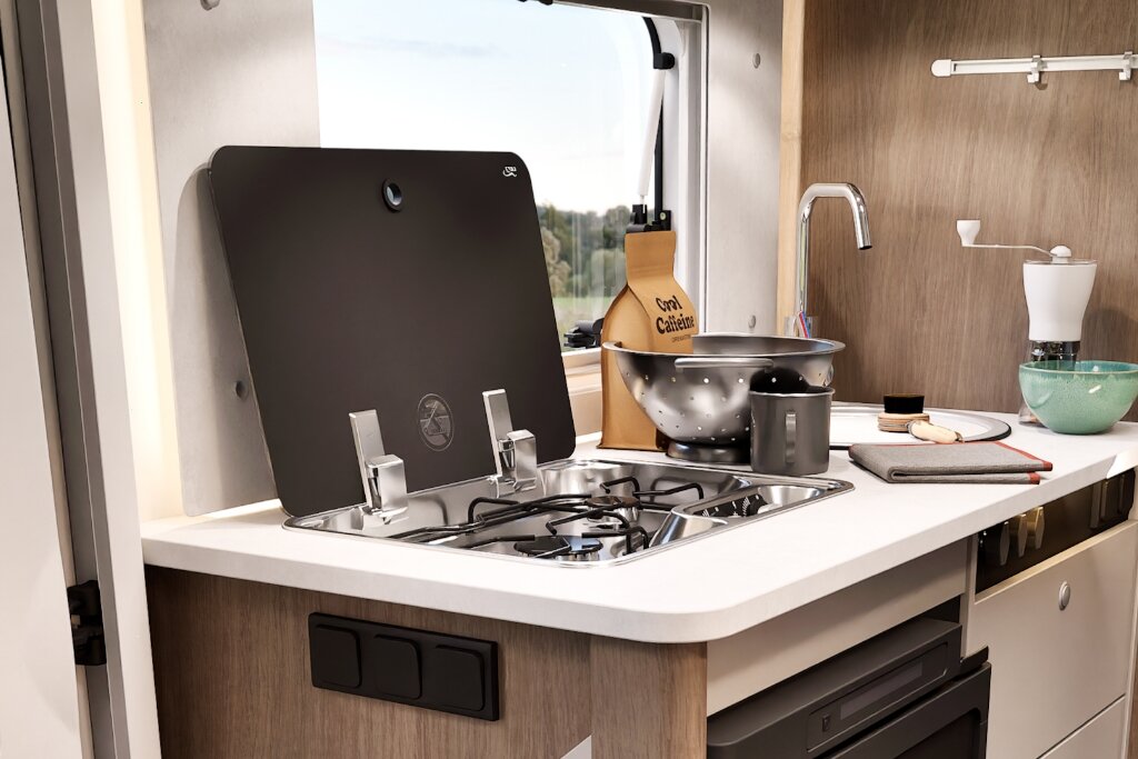 Küche im Carado Wohnmobil mit Gasherd, Spüle, Arbeitsfläche und Küchenutensilien am Fenster