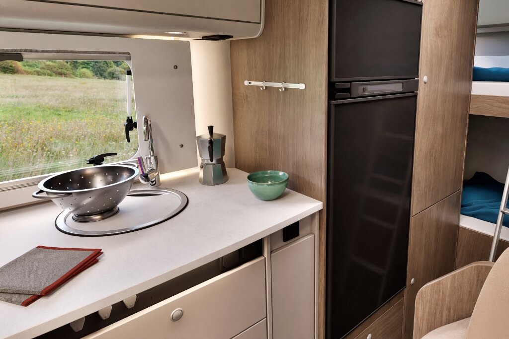 Küche im Carado Wohnmobil mit Spüle, Arbeitsfläche, hohem Kühlschrank und Blick auf Etagenbett