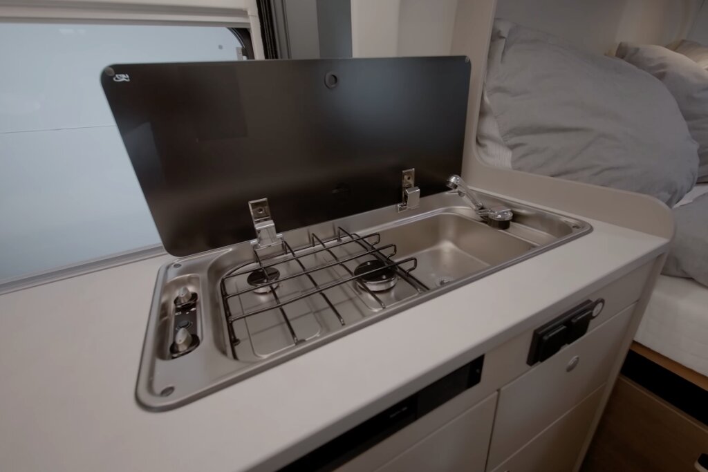 Küche im Carado Wohnmobil mit zweiflammigem Gasherd, Spüle und Glasabdeckung neben Bett
