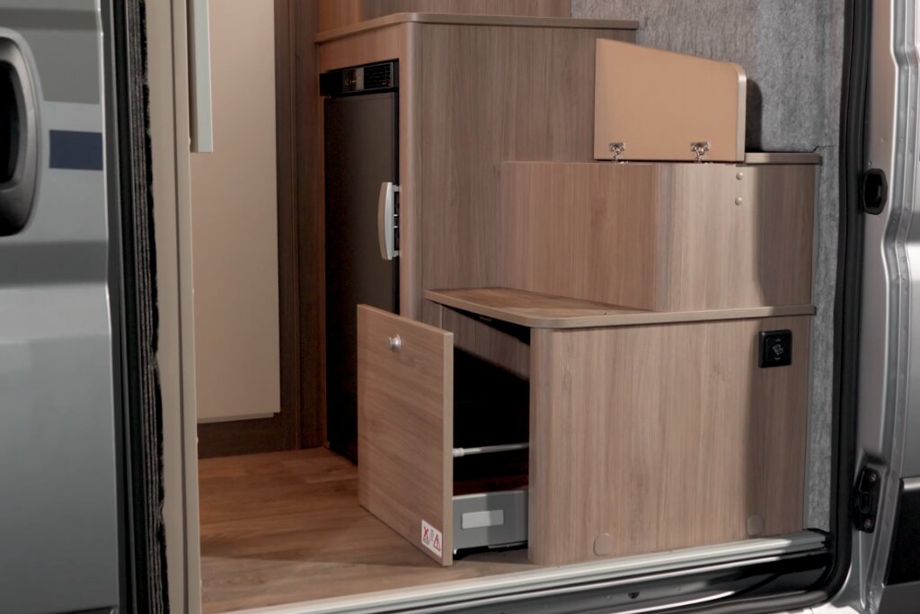Innenraum Carado Wohnmobil mit geöffneter Schublade, Kühlschrank und Holzschrank an der Seitentür