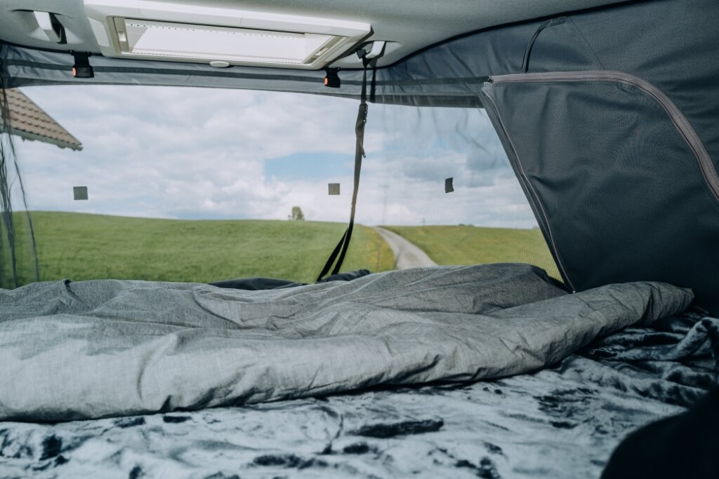 Schlafbereich im Carado Wohnmobil mit aufgeklappter Panoramazeltöffnung und Blick auf Feldweg und Wiese