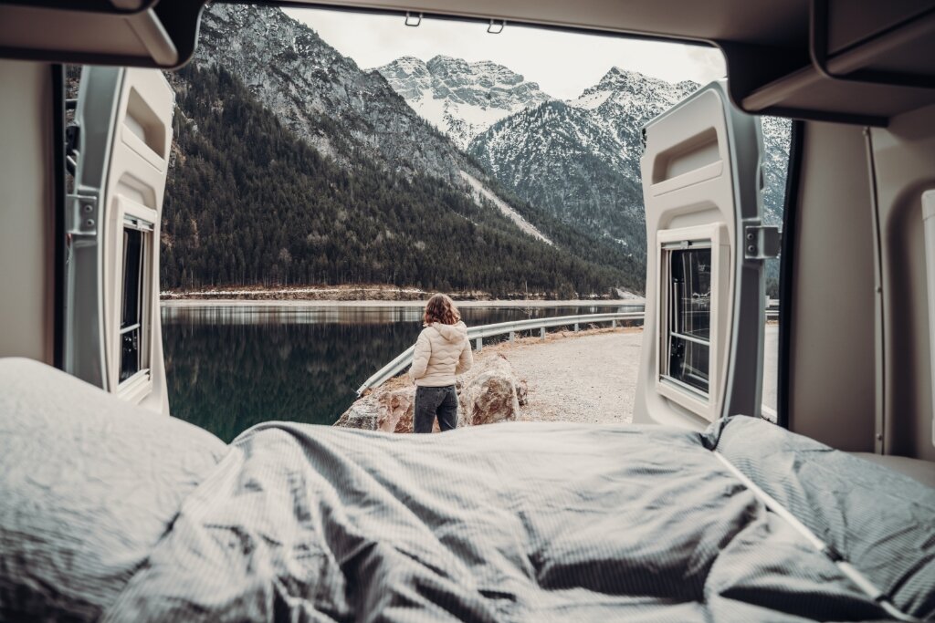 Blick aus dem Carado Wohnmobilbett auf Person am Bergsee vor schneebedeckten Bergen
