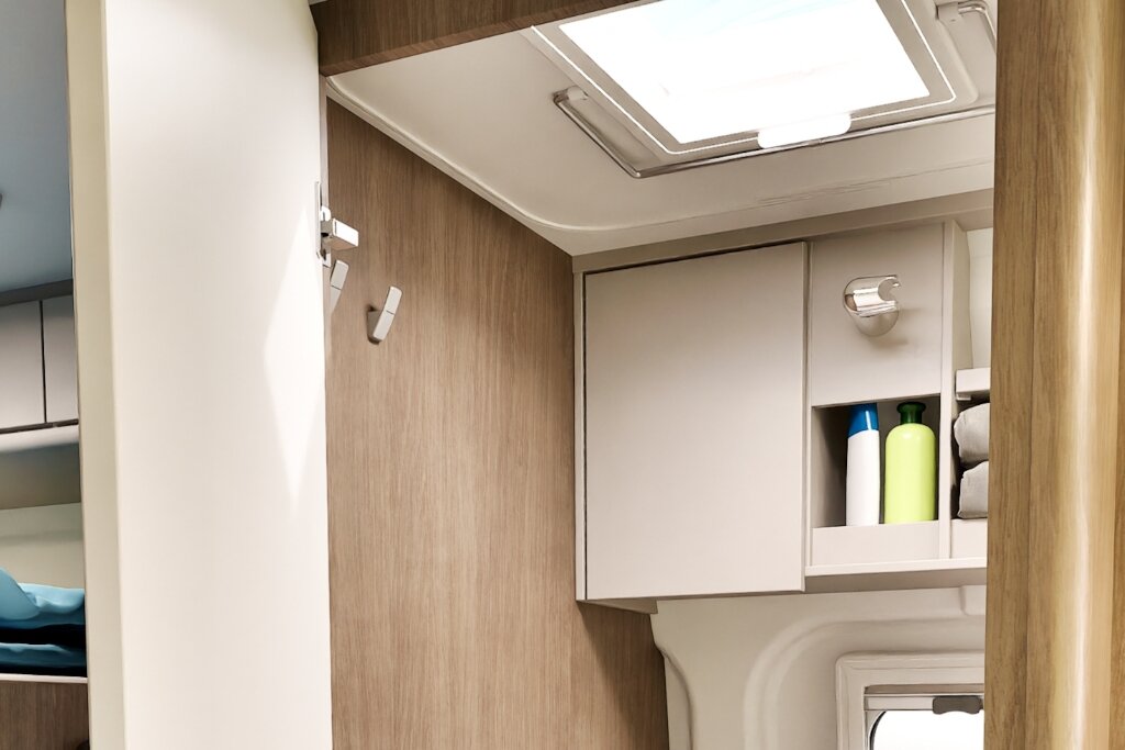 Badezimmer im Carado Wohnmobil mit Dachhaube, Hängeschrank und offenem Regal mit Flaschen und Handtüchern