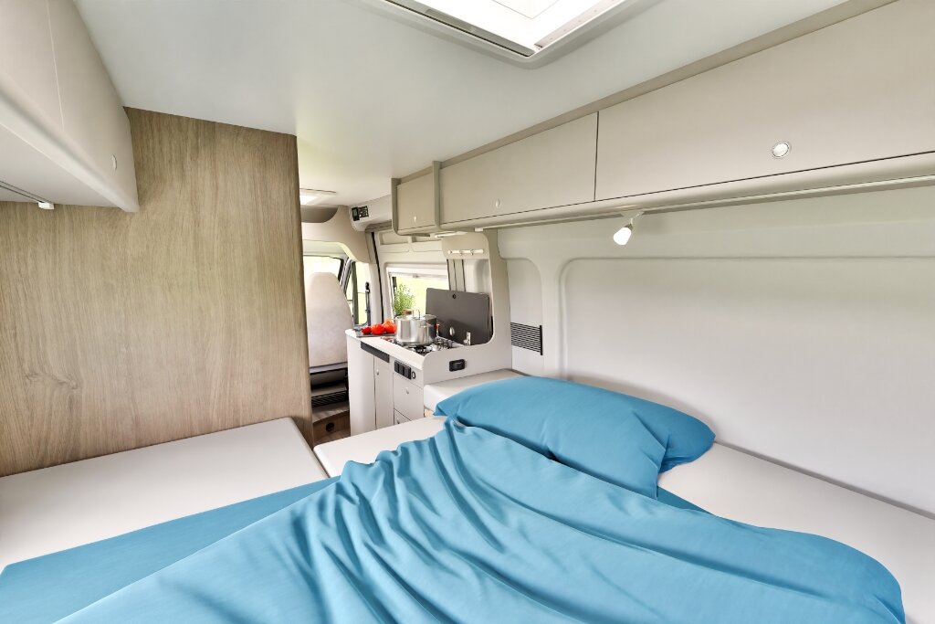 Schlafbereich im Carado Wohnmobil mit blau bezogenem Bett, Dachfenster und Blick zur kompakten Küche
