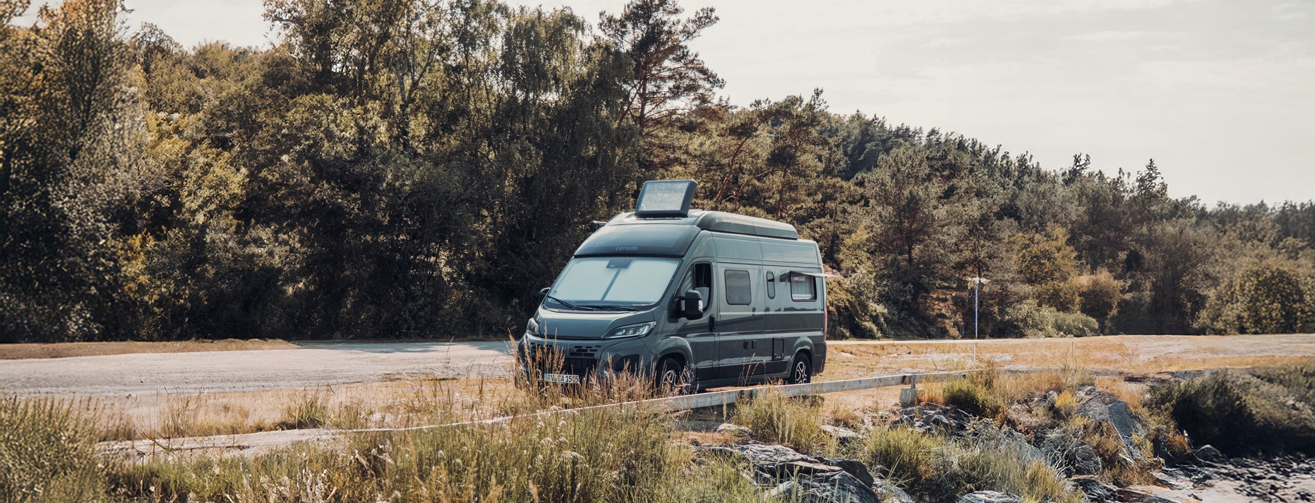 Carado-matkailuauto tien reunassa kallioisen rannan ja puiden lähellä, edestä ja sivulta nähtynä