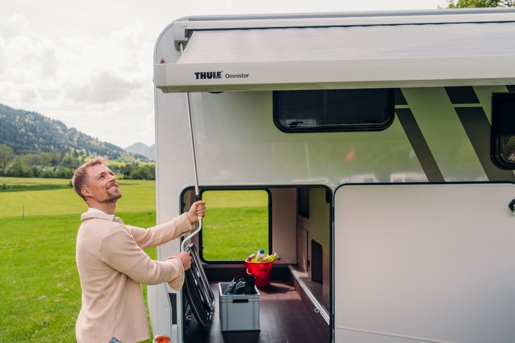 Person fährt Markise an einem Carado Wohnmobil aus, geöffnete Heckgarage auf Wiese vor Berglandschaft